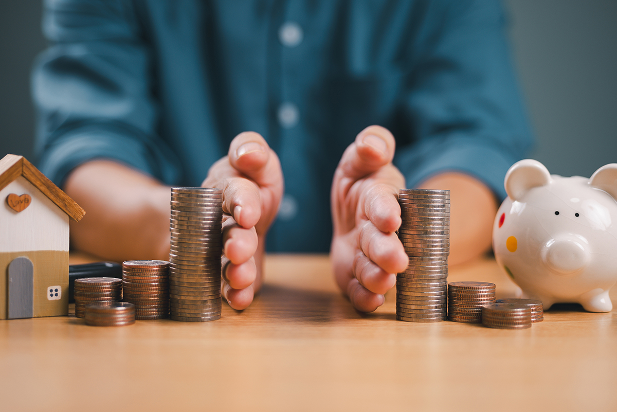 Stock photo of a man's hands dividing two nearly equal stacks of coins on a table, with a piggy bank next to the stacks of coins on the right, and a small wooden home next to the coins on the left.