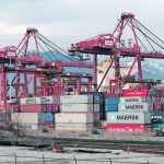Shipping containers sit stacked on the dock beneath large cranes with the mountains in the background at the Port of Vancouver.