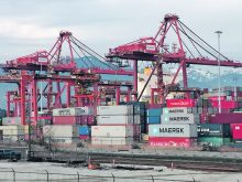 Shipping containers sit stacked on the dock beneath large cranes with the mountains in the background at the Port of Vancouver.
