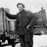 An archival photo from the 1930s of a man standing at the open-ended wooden box on the back of a truck next to a metal jug containing milk.