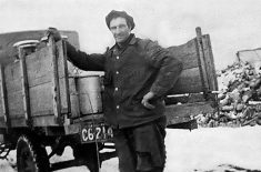 An archival photo from the 1930s of a man standing at the open-ended wooden box on the back of a truck next to a metal jug containing milk.