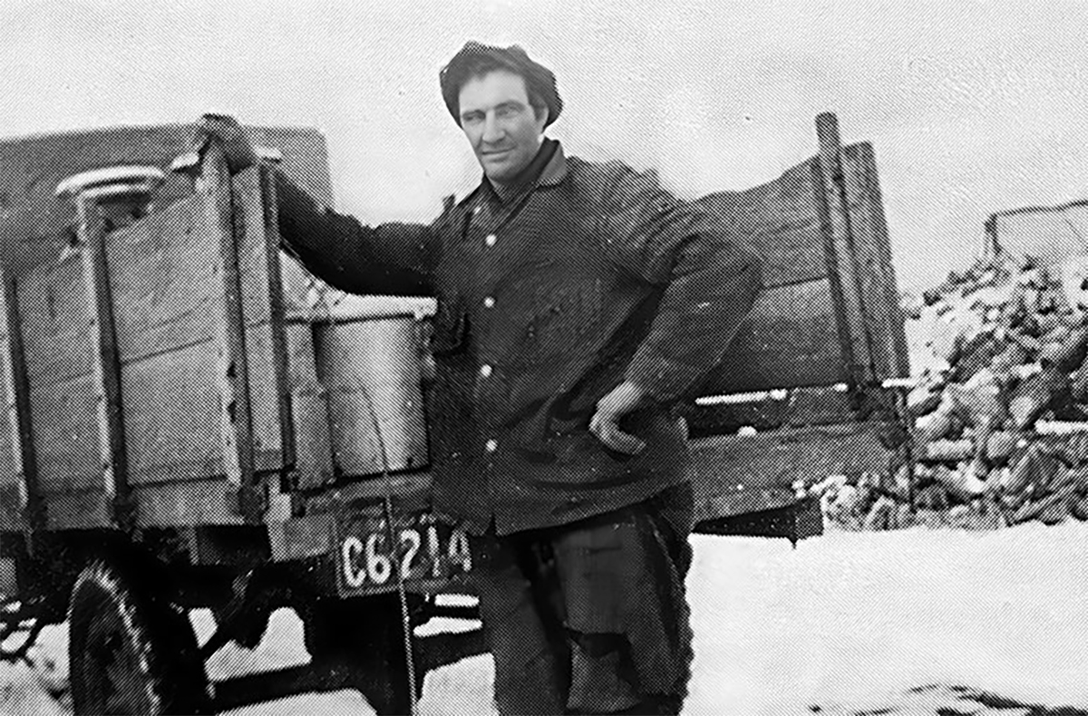 An archival photo from the 1930s of a man standing at the open-ended wooden box on the back of a truck next to a metal jug containing milk.