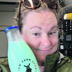 A woman smiles as she holds up a glass bottle of light blue milk next to her face.