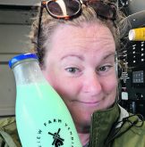 A woman smiles as she holds up a glass bottle of light blue milk next to her face.