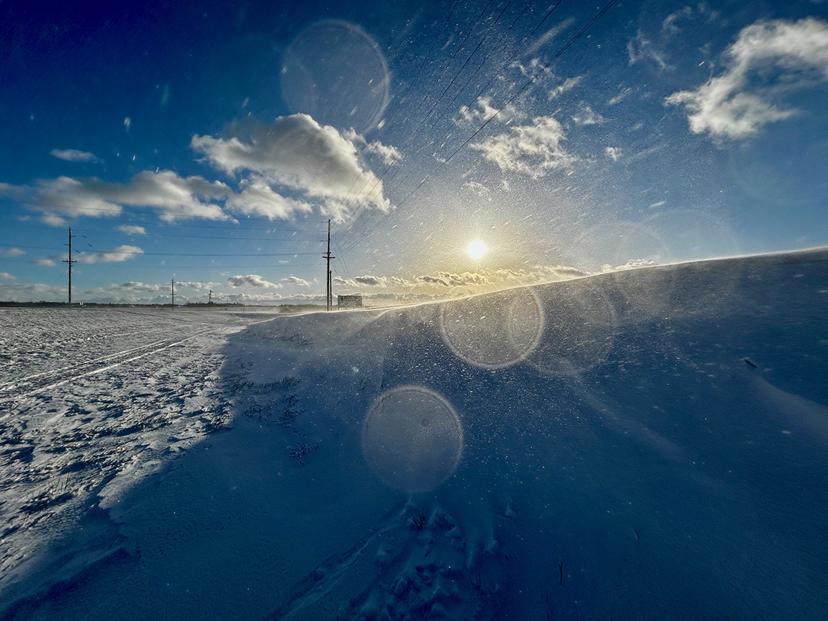A low angle shot of snow blowing off a snow drift on a cold day with a blue sky and the sun low to the horizon.