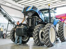 A man lifts the hood of a large, blue 8-tired New Holland tractor that is parked inside a shop.