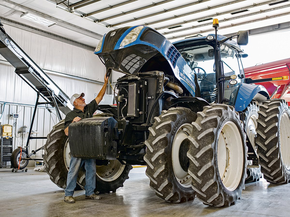A man lifts the hood of a large, blue 8-tired New Holland tractor that is parked inside a shop.