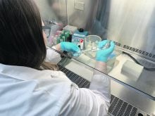 A woman waering a labcoat and blue latex gloves works under a ventilation hood in a lab.