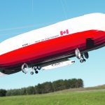 A cigar-shaped, red and white Buoyant Aircraft Systems International air ship takes off above a green field with some trees in the background.