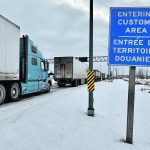 Semi truck are lined up on a highway in winter next to a blue sign that reads, "Entering Customs Area."