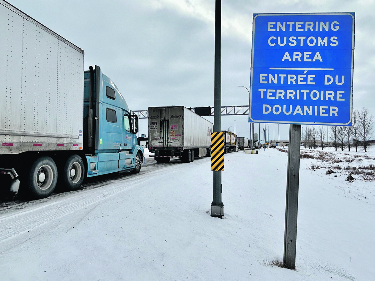 Semi truck are lined up on a highway in winter next to a blue sign that reads, "Entering Customs Area."