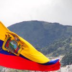 The flag of Ecuador ripples in the wind with low mountains visible in the background.