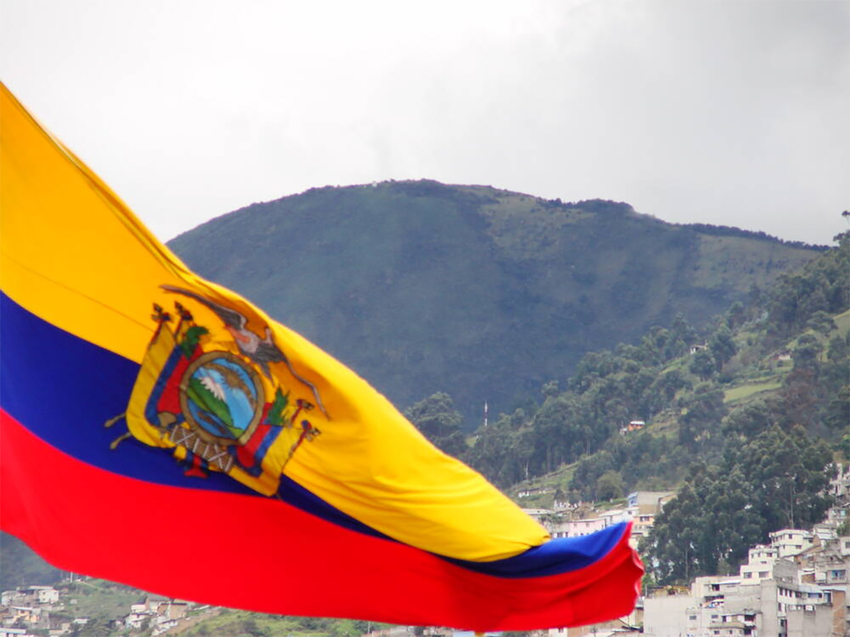 The flag of Ecuador ripples in the wind with low mountains visible in the background.