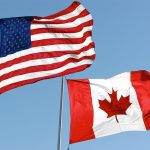American and Canadian flags fly side-by-side against a clear blue sky background.