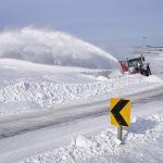 A tractor with a large, front-mounted snowblower throws snow high into the air as it clears a road.