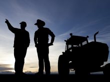 Two farmers talk at dusk next to a tractor, all perfectly silhouetted against the pink and orange glow coming from over the horizon.