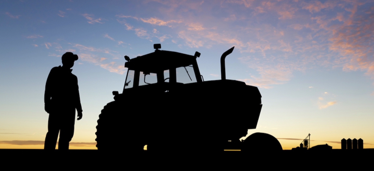 A farmer stands next to a tractor in a field, both perfectly silhouetted against the rising sun.