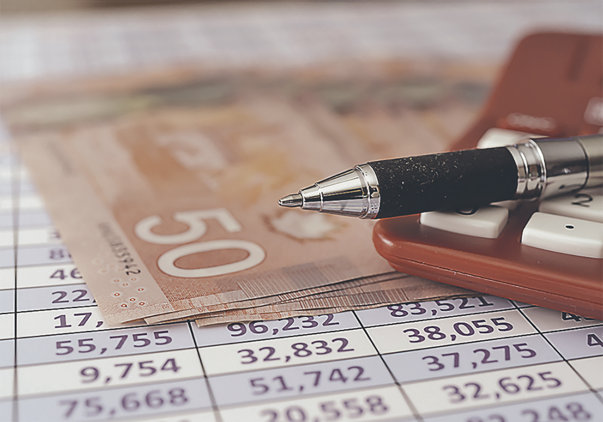 A pen rests on top of a calculator next to small number of $50 bills, all sitting on top of what looks like a spreadsheet filled with numbers.