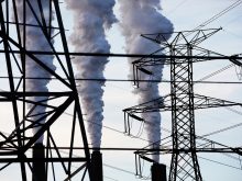 Steam billows from the stacks of a large industrial facility, with high-voltage electrical transmission towers and their power lines in the foreground.