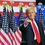 U.S. president Donald Trump stands, pointing directly at the camera with his right index finger, in front of a row of U.S. flags.