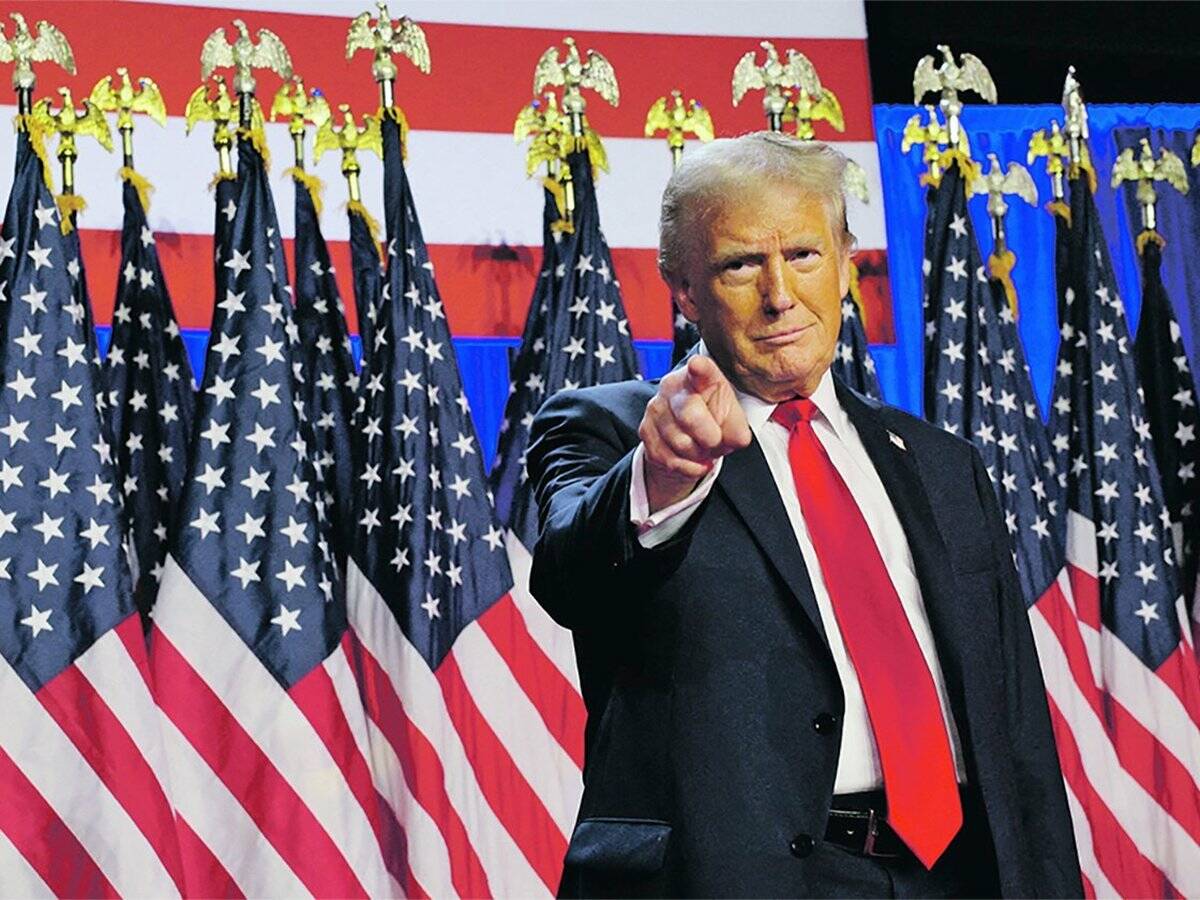U.S. president Donald Trump stands, pointing directly at the camera with his right index finger, in front of a row of U.S. flags.
