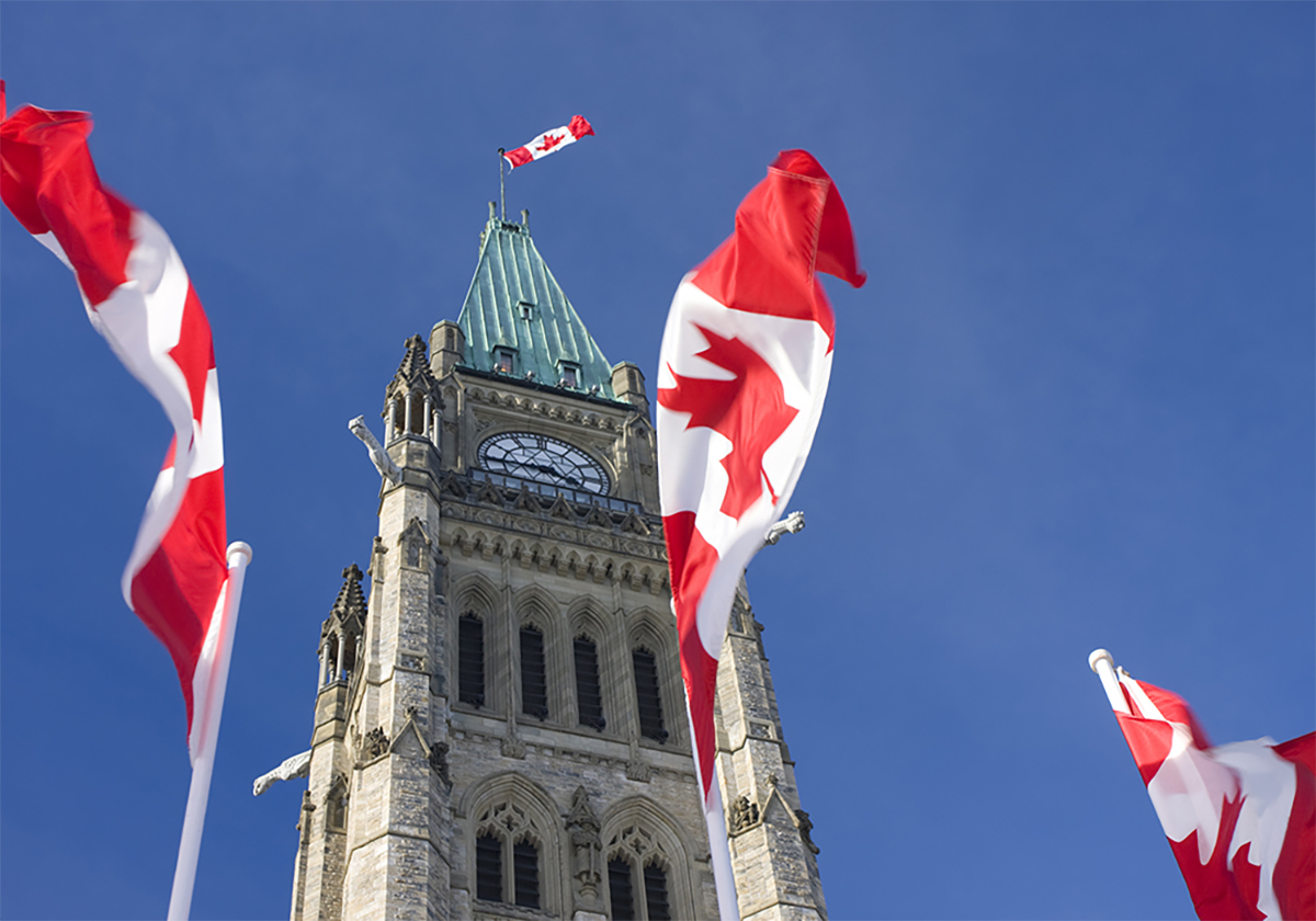Looking upward at the Peace Tower on Parliament Hill in Ottawa between three Canadian flags on poles on the ground.