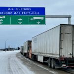 Trucks are lined up on a highway under as sign that reads, "U.S. Customs" as they wait to cross the border.