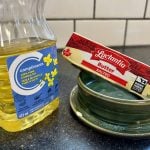 A stick of butter and a container of canola oil sit next to one another on a kitchen counter.