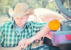 A stock photo of a young farmer in a cowboy hat leaning against his tractor and staring at the ground.