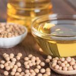 Soybeans sit in a bowl and some are spilled on a table top next to a bowl of seed oil.