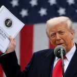U.S. president Donald Trump holds up a thick document entitled, "Foreign Trade Barriers" while speaking in front of a large American flag.