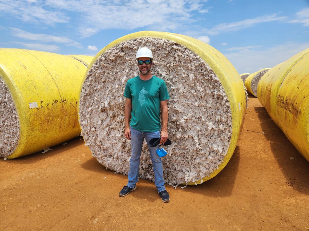A man stands in front of a large round bale of cotton wrapped in yellow plastic film.