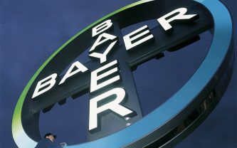 Looking up from the ground at a large, round Bayer logo sign with a man wiping it clean with a cloth at the bottom.