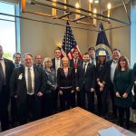A group of men and women in suits stand around a boardroom table with an American flag in the background.