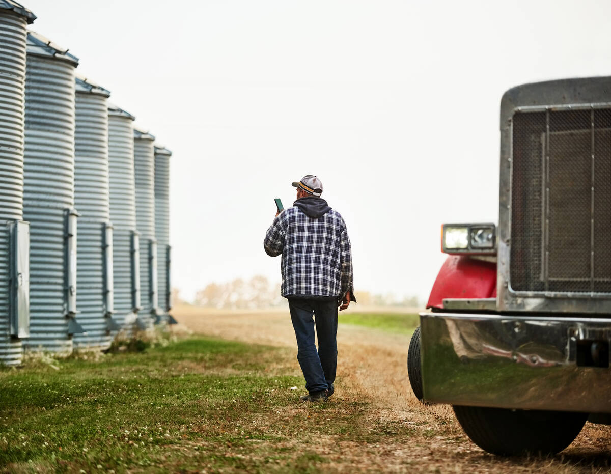 A farmer is talking on his smartphone as he walks in front of a line of steel grain bins.