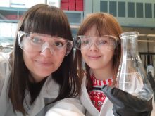 Heather Blewett and her daughter, Madelyn, wearing protective glasses, white lab coats and holding a flask in a lab.