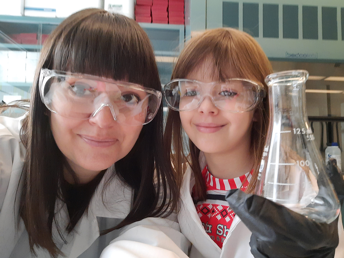 Heather Blewett and her daughter, Madelyn, wearing protective glasses, white lab coats and holding a flask in a lab.