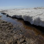 Water runs in a ditch next to some melting snow on a sunny day.