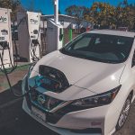 A Nissan Leaf EV sits at a charger station with the charging cable connected to the front of the vehicle.