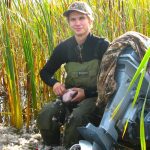 A young man sits on the edge of a boat on shore, next to its outboard motor, plucking a duck's feathers.