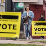 Yellow Elections Canada "Vote" signs are stuck in the grass while people walk toward them in the background.