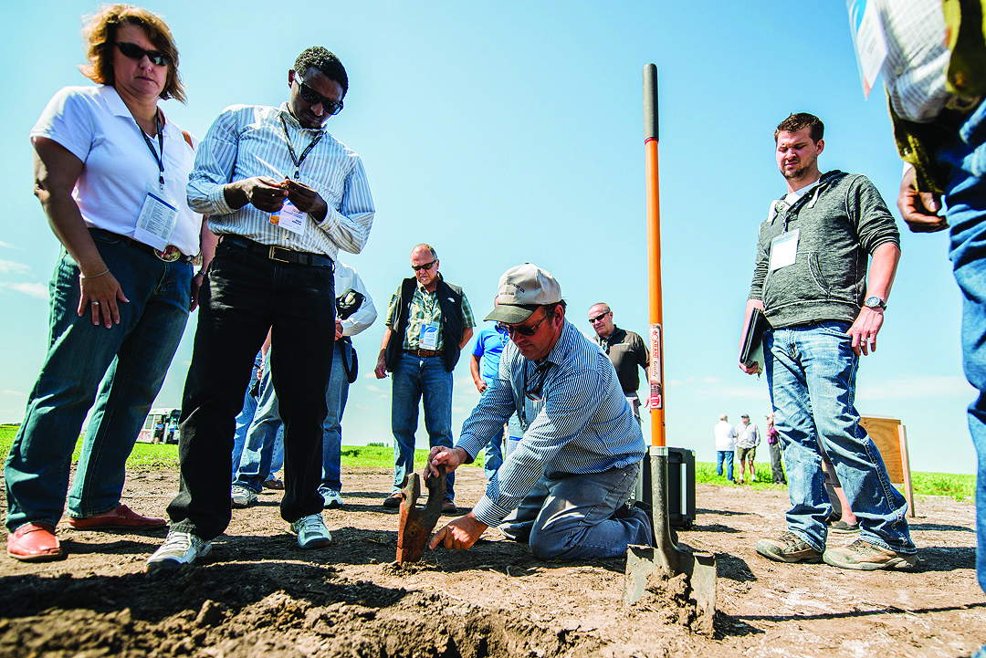 A group of people stand outdoors on a sunny day, watching a man kneeling on the ground as he digs into the soil with a small tool. Some people take notes or photos, suggesting a field demonstration or workshop.