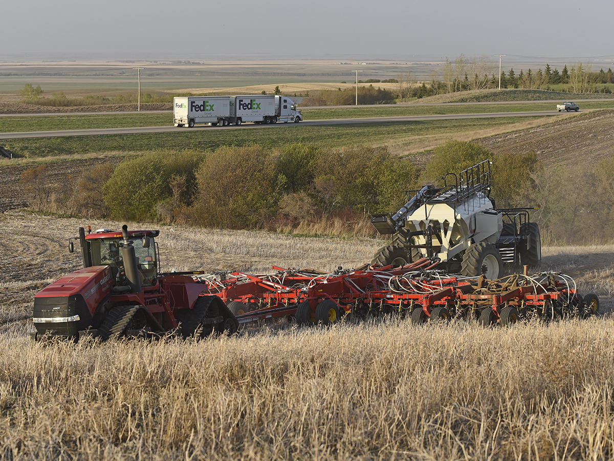 A large, red, tracked tractor pulls an air seeder rig in some hills next to a highway with traffic on it in the background.