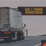 A semi truck moves down a highway about to pass beneath a sign that reads, "USA | Canada Border Ahead."
