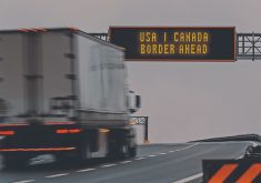 A semi truck moves down a highway about to pass beneath a sign that reads, "USA | Canada Border Ahead."