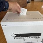 A man's hand deposits a ballot into a cardboard "Elections Canada" ballot box during voting in the 2025 federal election.