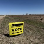 A yellow Elections Canada "Vote" sign is stuck in the ground alongside a grid road in rural Saskatchewan.
