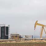 A crude oil pump jack stands in a field next to a couple of large black oil holding tanks.