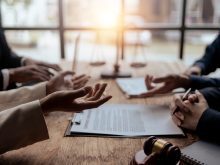 The animated hands of people seated on opposite sides of a boardroom table with a gavel in the foreground, and a blurry "scales of justice" statue in the background.