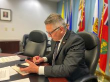 Newly-appointed Minister of Agriculture Heath MacDonald is seated at a desk with his phone in his left hand and writing notes with his right hand.